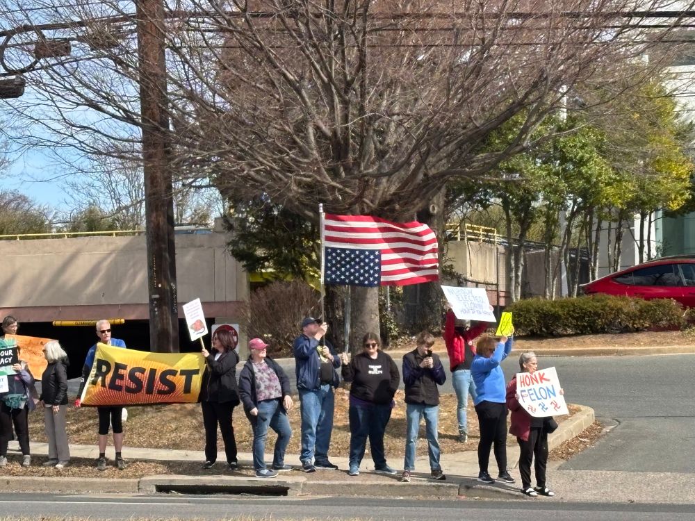 Crowd of protesters in front of Tesla dealership with American flags upside down, resist and other anti-musk signs. My favorite was “This machine funds facists.”