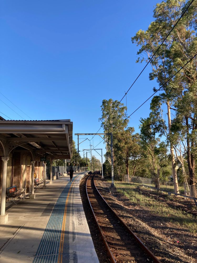 Windsor railway station platform on a clear day, with a person walking along the platform. The view includes tracks curving to the right with trees to the right of the track. A clear blue sky can be seen above the station and trees.