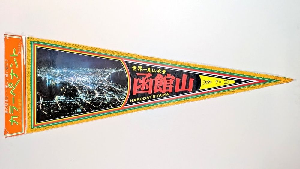 1, 函館山のペナント。紙に写真をプリント。黄色いフリンジ装飾付き。旗はオリジナルの透明ビニル袋入り、紙のタグ付き。タグのタイトルは "カラーペナント, 旅の記念に···美しい想い出に!!, あなたのお部屋に!" (橙, 黄緑, 白) 。ブランド名は "オクモト" (白) 。