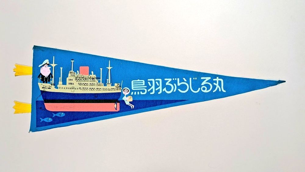 1, 鳥羽ぶらじる丸のペナント。青い布にイラストをプリント。旗の左にリボン形の黄色い装飾2つ付き。