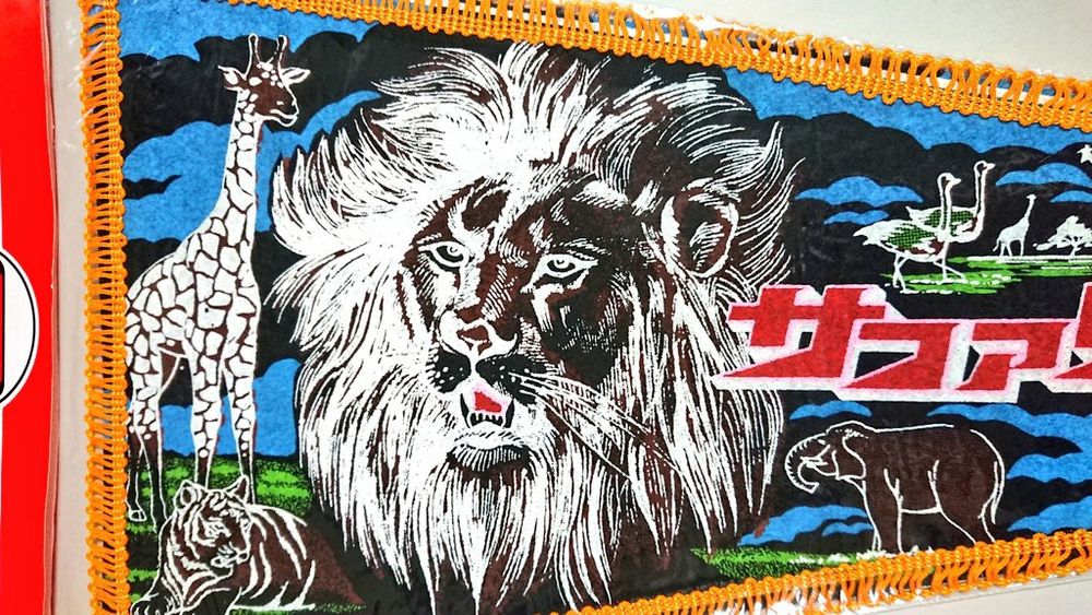 3, Enlargement of the illustration. The head of a male lion with a magnificent mane is depicted in the center. Giraffes, elephants, tigers, and ostriches are depicted against a spacious savanna landscape.