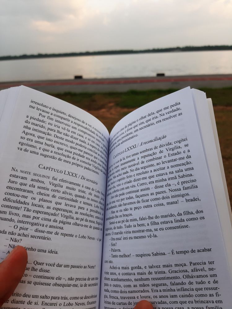No primeiro plano um livro aberto atrás é possível ver uma orla, um rio e árvores na outra margem.