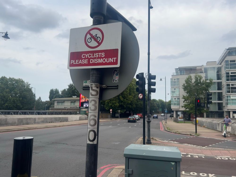No-cycling sign with text “Cyclists please dismount”, four lane urban road in the background.