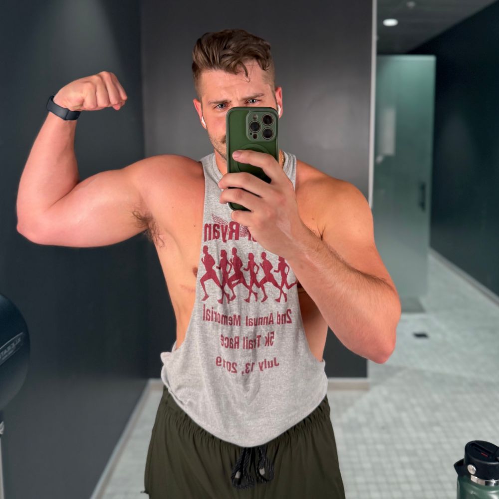 Mirror selfie of Adam (Lemonboy) flexing in a locker room wearing a gray tank and dark olive green shorts.