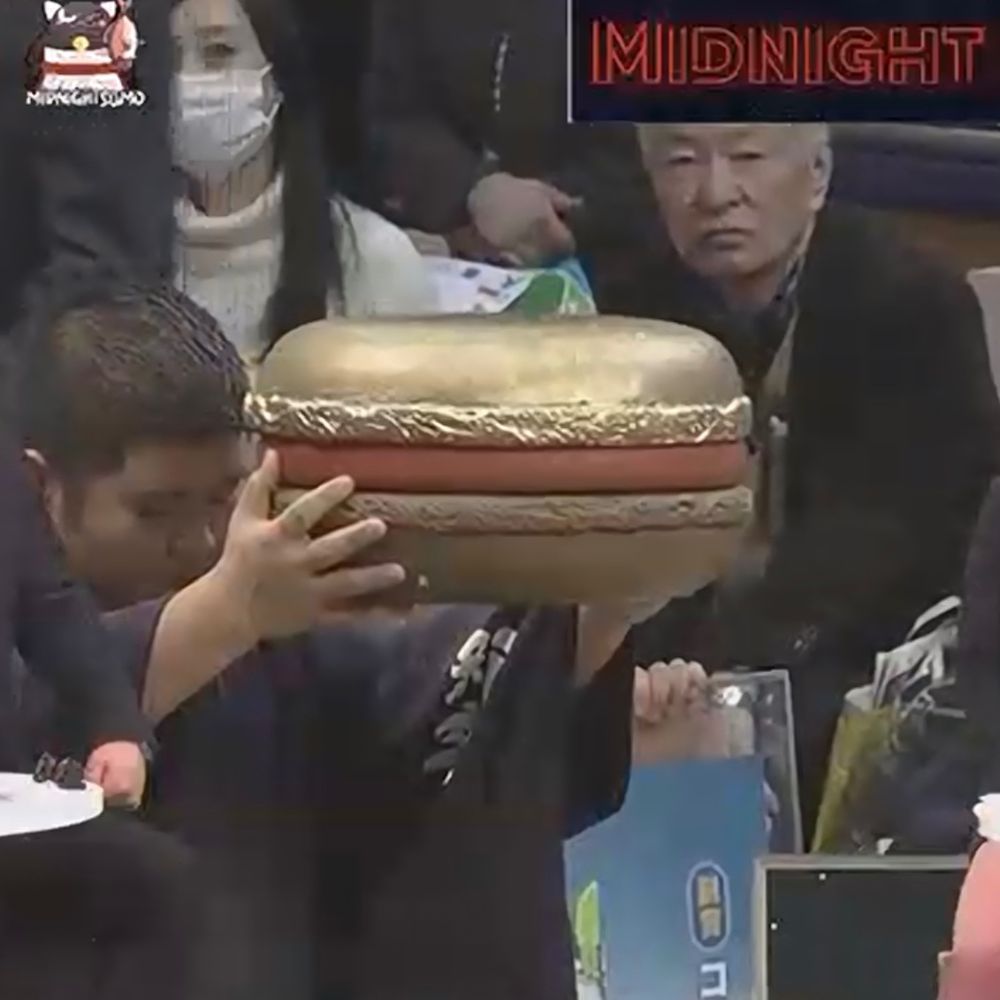 A screenshot of a man holding up a giant, golden macaron at the end of a sumo tournament.