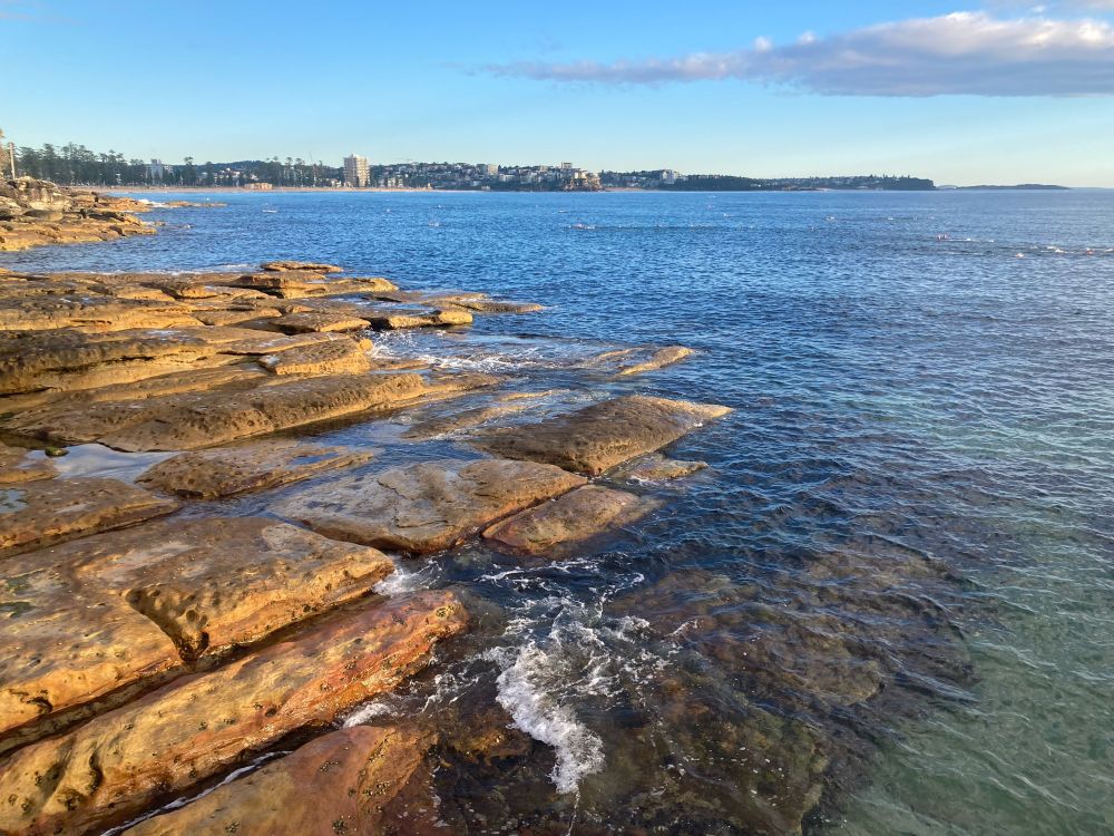 Clear sea water with low waves washing onto orange brown square shaped rocks on bottom left. A long beach with trees behind it in the distance 