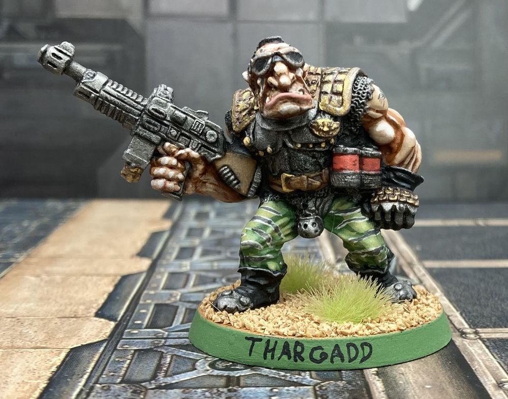 A Warhammer miniature of an ogre, wearing camo pants and a metal breastplate, holding a machinegun. Written on his base is his name: THARGADD.