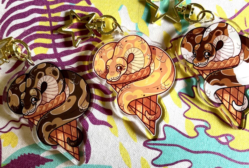 Three ball python acrylic keychains with a star clasp on a brightly colored background. From left to right they are regular ball python, banana ball python, and pied ball python