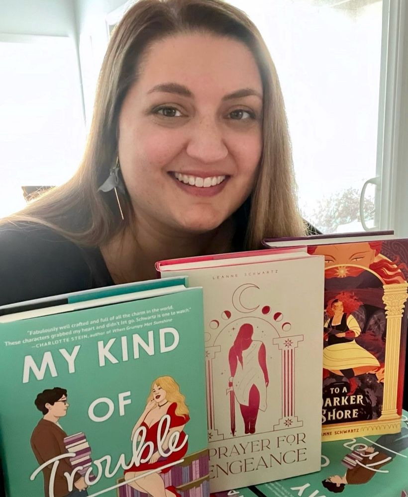 Me, a fat white person, smiling and sitting and leaning over my three books standing on display: My Kind of Trouble, A Prayer for Vengeance, and To a Darker Shore, with soft light from a two windows behind me

My Kind of Trouble book cover: teal background with a bold white san serif font reading MY KIND OF across the top and between two figures, TROUBLE written in large script over the two figures, and a small script font reading “a novel” tucked into a bookshelf near the bottom. The two figures are a tall dark-haired man in glasses, brown tweed jacket, jeans, and sneakers, carrying an overly tall stack of books and smiling at a glamorous fat blonde woman in a low-cut red dress, choker, and red lipstick sitting on a low bookcase, holding a book in her lap, and smiling back slyly. The books are in pinks and purples. The author L. A. Schwartz is written at the bottom.

A PRAYER FOR VENGEANCE cover, reading “Leanne Schwartz, A PRAYER FOR VENGEANCE” and featuring a fat teenage girl in gladiator sandals and a sparkling draped dress with a high slit revealing one thick thigh as she stalks forward, lowered sword ready in one hand and dripping three drops of blood. She is surrounded by classical columns topped with stars and an archway of moon phases, topped with a large crescent moon. The cover is in a pink to dark wine color scheme, on a parchment background.

A book cover for TO A DARKER SHORE featuring a fat girl in a bodice and skirt running and carrying a crossbow, a wry, determined smile on her white face looking over a shoulder, dark hair flying behind her. She’s above the pit of hell and surrounded by levels of hell, with fire, devils, mirrors, and towers rising up to mountains and a sun in the sky, watched over by a large face of a golden god. The image is set between two golden columns. A sad monster boy watches the girl from behind a broken mirror, bloody hand pressed to the glass. Colors are red, gold, and purple. Author’s name at the bottom: Leanne Schwartz