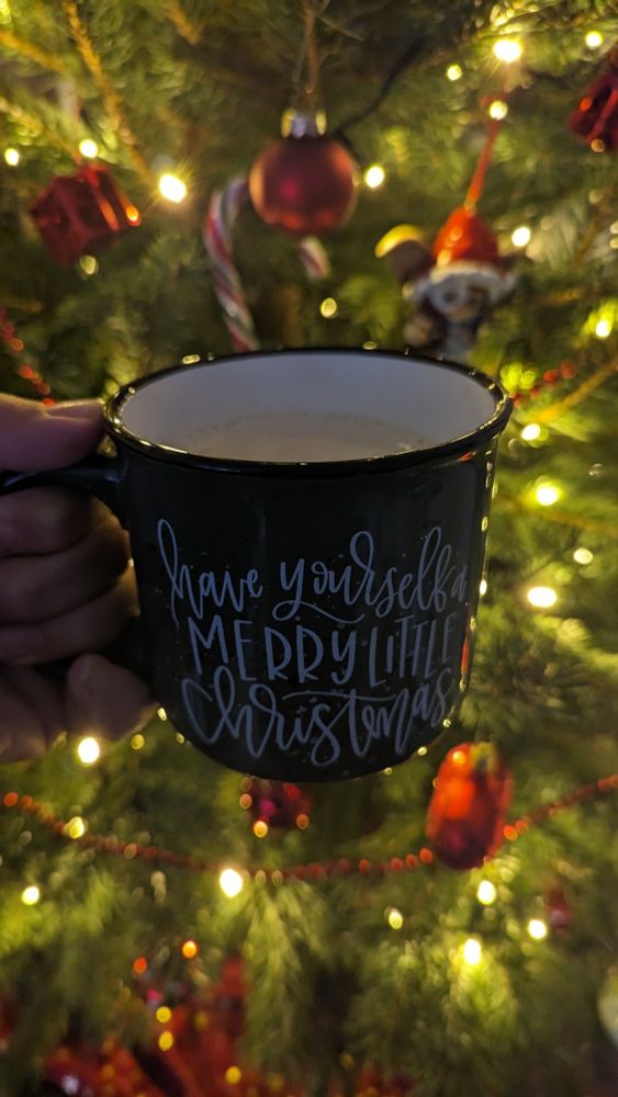 Im Vordergrund eine Tasse Kaffee, Tasse ist dunkelgrün und trägt den Schriftzug "Have yourself a merry little Christmas". Im Hintergrund eine Tanne, geschmückt mit roten und weißen Kugeln, Zuckerstangen und Gremlins.