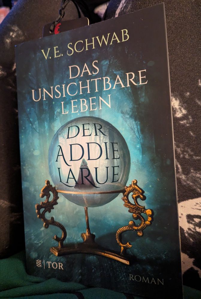 Der Roman "Das unsichtbare Leben der Addie LaRue" von V.E.Schwab. Das Cover ist smaragdgrün und zeigt einen angedeuteten Wald. im Vordergrund steht eine Kristallkugel? Ein Stundenglas ? in dem man eine Person, gehüllt in ein Cape mit Kapuze von hinten sieht