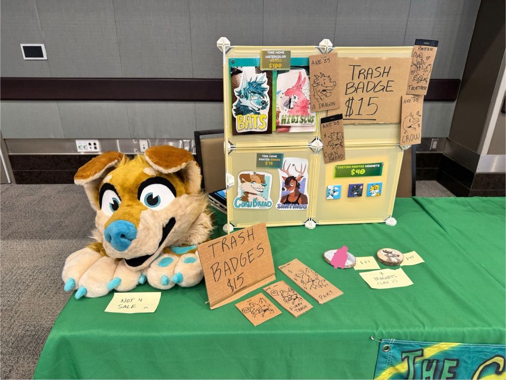 Display stand showing signs that say “Trash Badges $15” along with examples of small, phone-sized cut pieces of cardboard with different animals on them. There is also a golden retriever fursuit head and several art pieces on display as examples of other badge types.