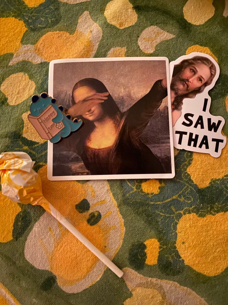 A dumdum lollipop, an enamel pin of a dinosaur reading a book called “happy times” and two stickers, the Mona Lisa dabbing, and Jesus said “I saw that.” His tattooist is adorable. 