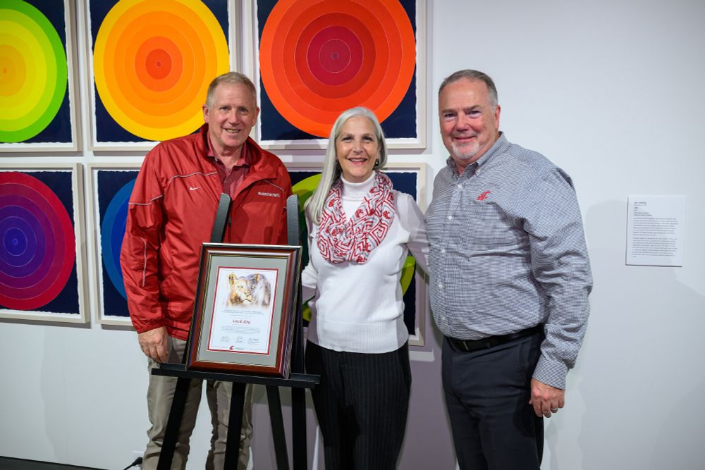 Three people pose in front of colorful artwork with a framed certificate on an easel in front of them
