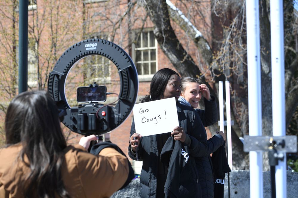 Two WSU students being filmed on campus, smiling, and standing back-to-back while one of them holds a sign that reads: "Go Cougs!"