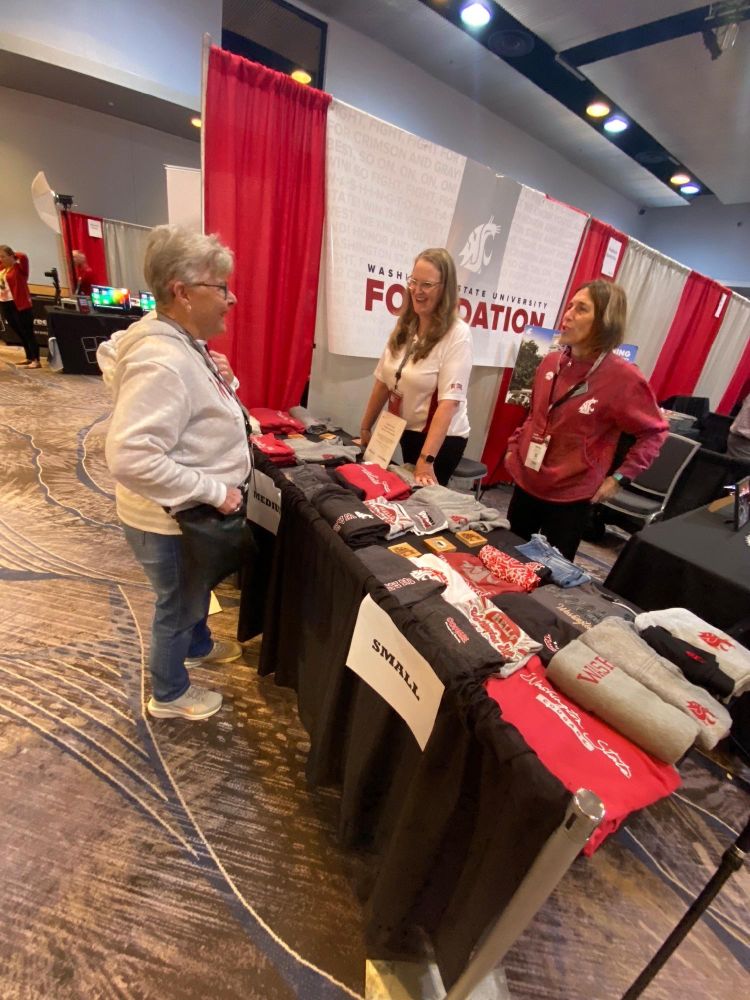 Three individuals at a Washington State University Foundation booth discussing over a table displaying merchandise.