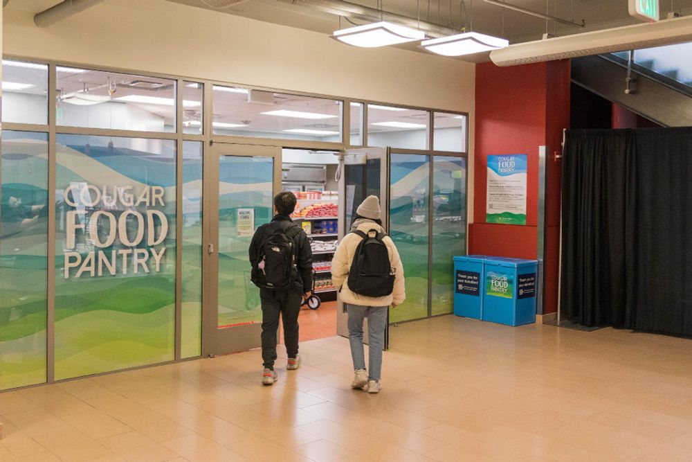 Two people with backpacks walking towards the entrance of the Cougar Food Pantry in a building.