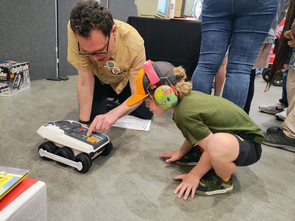Dr James Sumner and a young boy with colourful headphones interacting with a robot toy at Dr Sumner's Electric Dreams stall. Photo by Dr Erin Beeston.