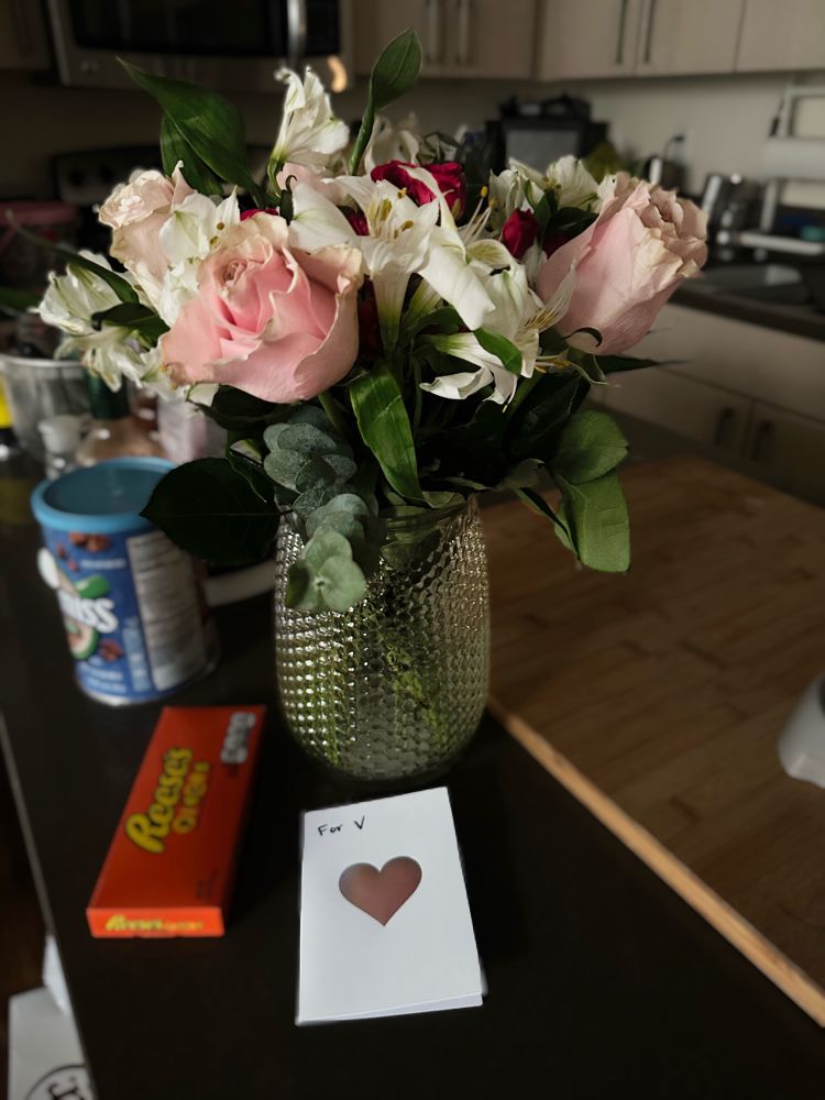 A bouquet of flowers in a vase on a kitchen island. There’s a card in front of them with a holographic heart on it and the handwritten words “For V.” There’s a box of Reese’s pieces beside it. 