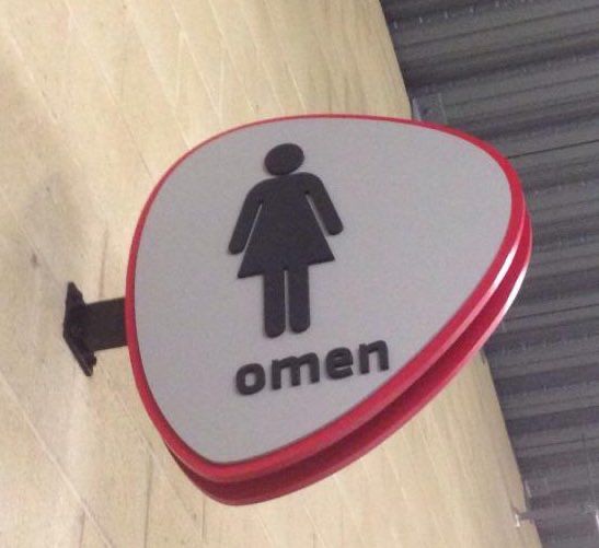 Image of a red, black, and white women's bathroom sign mounted on a tan tile wall, it is missing the letter 'W.' Without the W, it spells "omen."