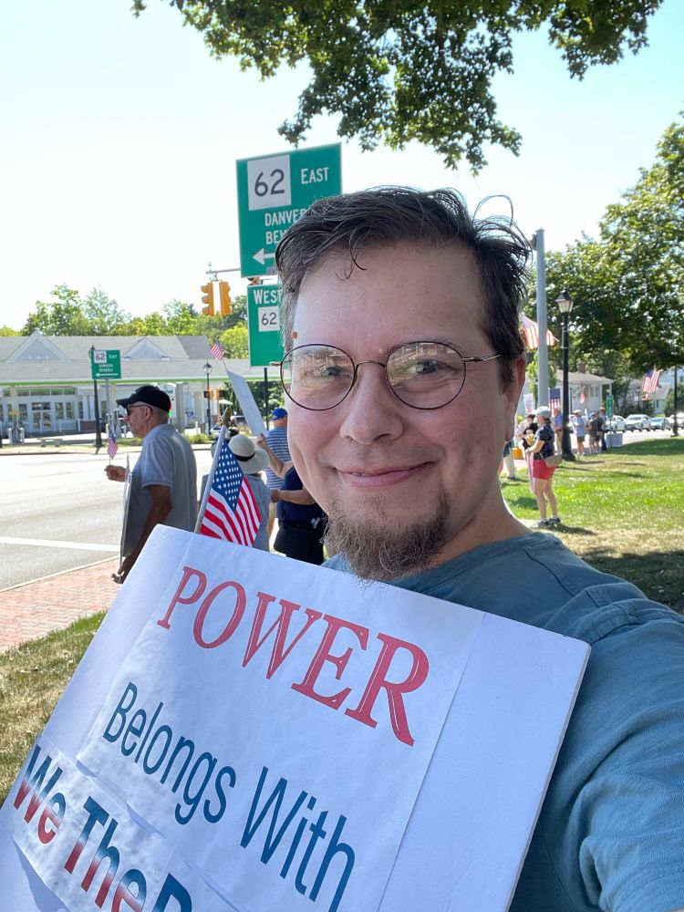 Beth Andres-Beck smiles at the camera. They have short hair, a goatee and glasses, and are holding a sign that says "Power Belongs With We The People". Behind them a line of people, many in ball caps and shorts and holding signs, stand along the sidewalk on a sunny day.