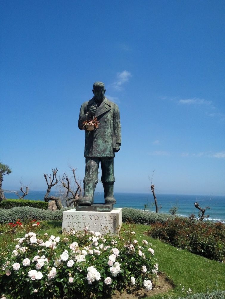 Escultura en piedra, cuerpo entero sobre pedestal en zona ajardinada.
Escultura del poeta José del Río Sáinz. Poeta del mar. 
Conocido popularmente como "El botas" por las grandes botas que lleva.
Situado en una zona ajardinada. 
Al fondo, el mar Cantábrico.