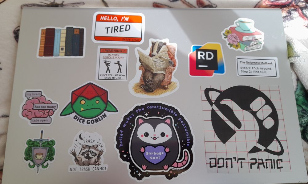 A grey laptop with several stickers.  They include references to Hitchhiker's Guide, Dice Goblins, Legends & Lattes, Trash Animals & dry humor.