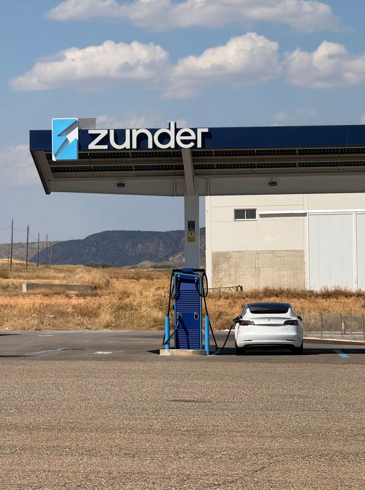 Un coche blanco cargando en una estación Zunder techada y en una zona de la meseta. 