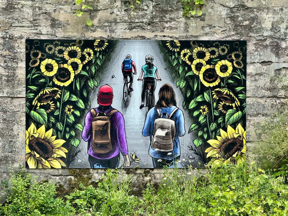 Caledon Trailway mural by Blaze Wiradharma inspired by the history of the area shows people walking and biking on the trails, surrounded by sunflowers