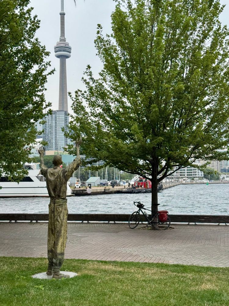 A sculpture of a man who came to Toronto during the Irish famine with trees and Toronto skyline in the background. The image is taken at Ireland Park.