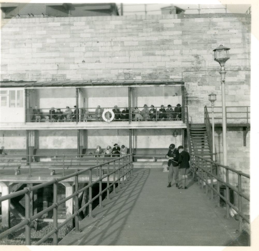 Unidentified photo taken around 1953 on the cost showing a two storey structure against a high stone wall with open front, forming a veranda with balcony above, both with bench seats and people sitting in them. 