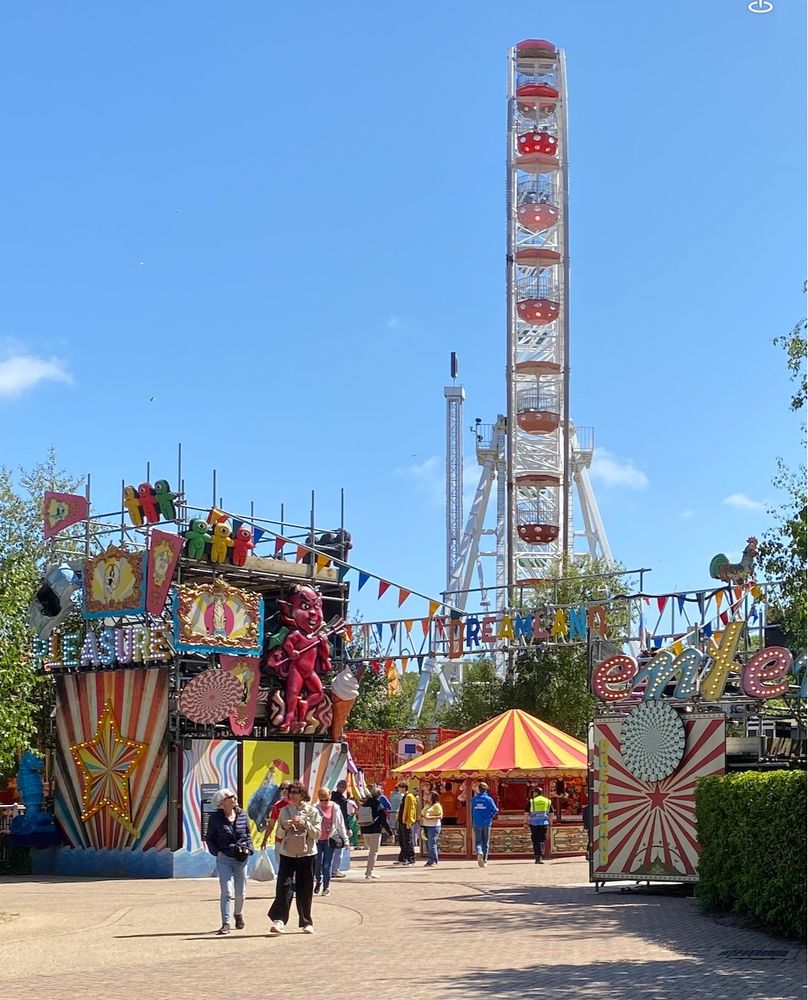 entrance to the funfair at Dreamland, with the big wheel in the background, the entrance is framed by two stands, the left one taller and with a jumble of signage and ornament including a large red plastic devil. There's a stall just past the entrance with red and yellow striped canopy over. Lots of bright primary colours.