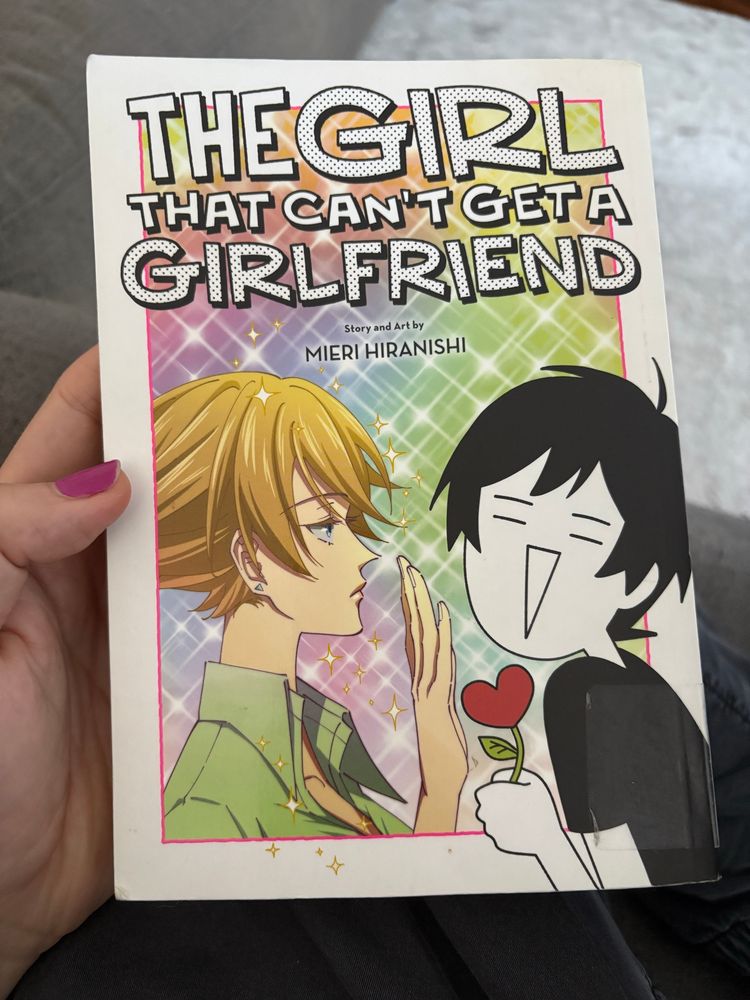 Photo of my holding the book The Girl That Can’t Get a Girlfriend by Mieri Hiranishi. The art shows a pretty shoujo-looking woman in colour holding hand up, blocking the black and white simpler drawing of the author holder a heart on a flower stem. The background is sparkly and colourful.