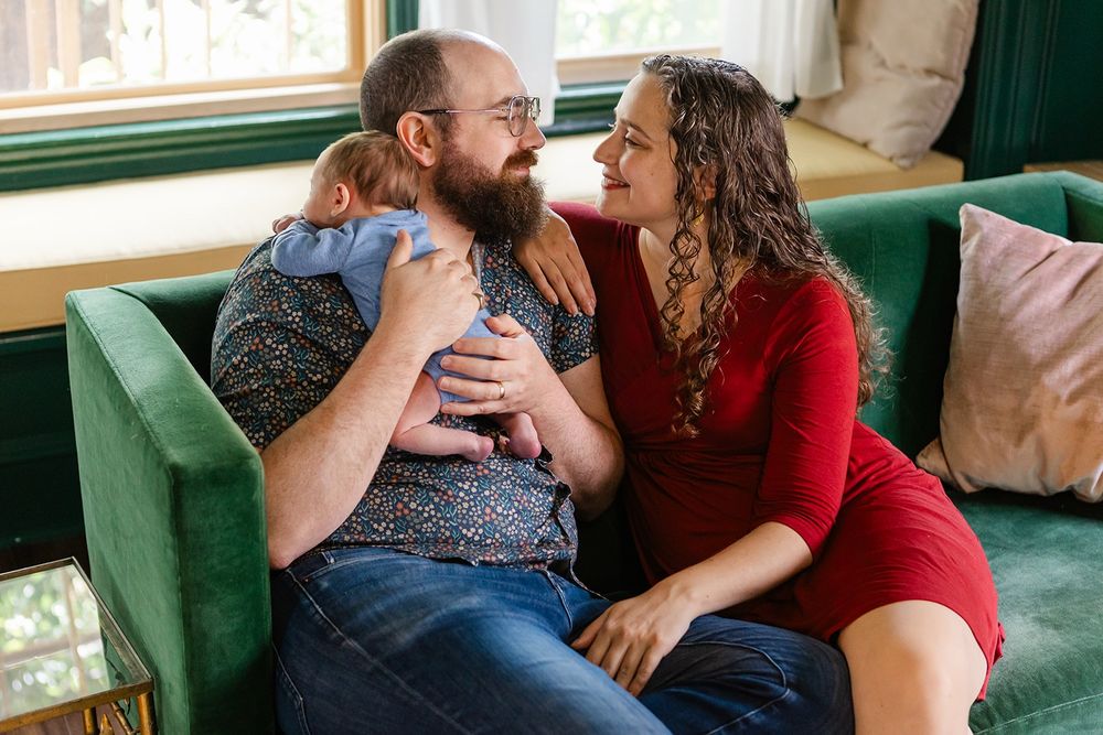 Portrait of Theo and Alana, sitting on a green velvet couch, holding a small baby in a blue onesie over his shoulder.
