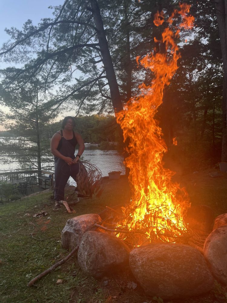 The fire is going - probably about 2m tall flames (taller than Steve). He is wearing a tank and linen overalls (one side undone). 