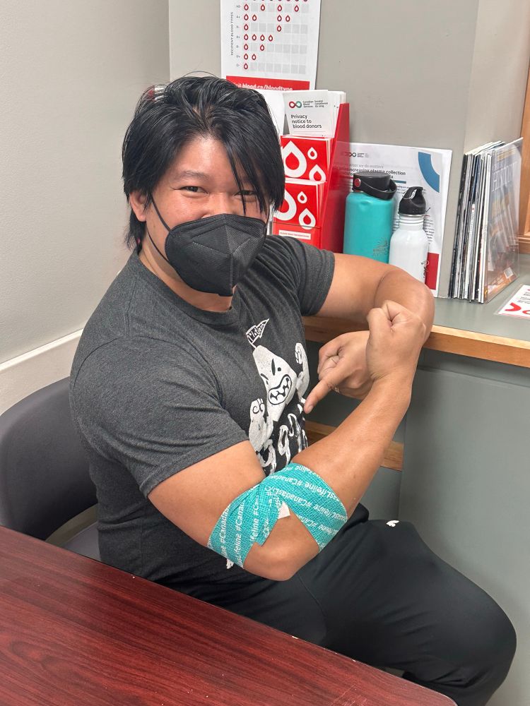 Steve sitting down after a blood donation. He’s in a black tshirt and black athleisure pants. He’s pointing at a teal wrap around his donation site. He has a KN95 mask on. 