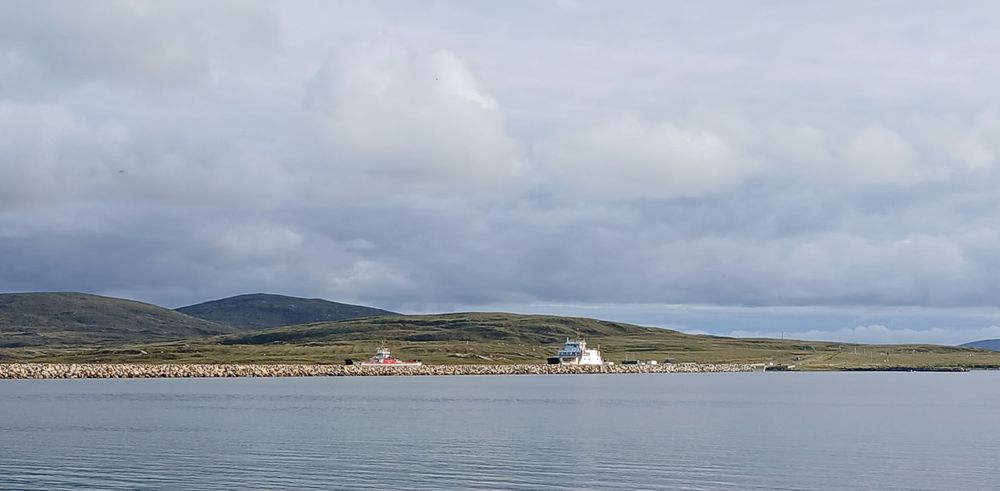 Two tiny ferries docked behind a causeway in the outer hebrides with low green hills and fluffy clouds behind