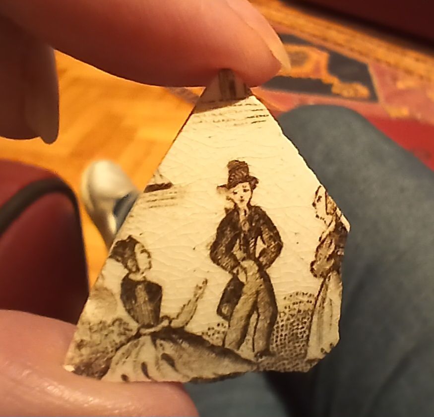 Small triangular fragment of pottery. Part of a boat in the backgound, on the sea with a possible tower on a small island behind. On land in foreground are a trio. Center is a gentleman in tails and tall hat. To his right a kneeling woman beseeching, although her arm is growing out of her pelvis. To his left a matron in a cap, arms folded, looking furious.
'Look, ye numpty Frankenstein, I'll gie ye DOCTOR. Ye said you could gie her a new arm. Look whit ye've made!'
