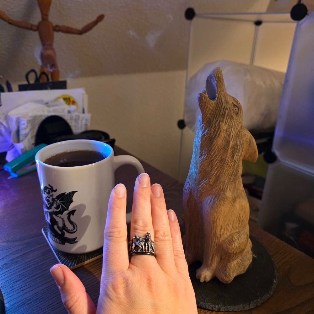A photograph showing a mug with the WorldAnvil crest, a wooden wolf whose mouth emits wisps of smoke, and a hand with a ring that shows five wolves.
