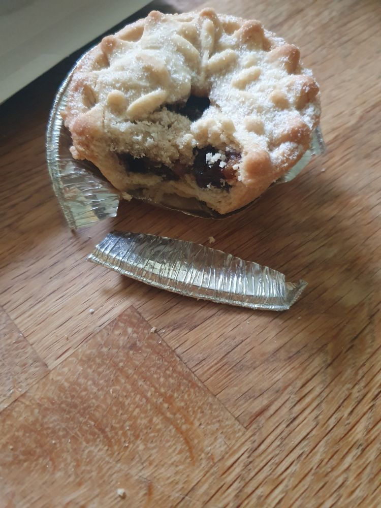 A mince pie in a tin with a bite taken out but the notable thing is the side of the tin has been completely torn off and is in pieces rather than daintily spread open for ease of biting 