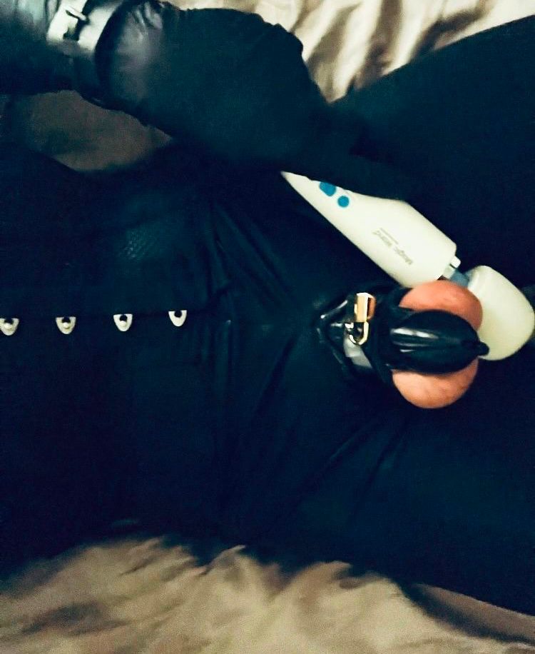 A white submissive male wearing a shiny spandex catsuit (unzipped at the crotch), mesh corset, black rubber gloves, leather wrist cuffs and shiny black silicone chastity cage (locked with a golden padlock) places a the head of a Magic Wand vibrator behind his achingly full and exposed testicles. The slow and methodical process of dressing in this tightly strapped, zipped, clasped, locked and laced has long since made him continuously drip with arousal. He yearns to have his confined cock free to be swollen and stroked, but he knows that forcing himself to cum inside the cage will be both intense and ruined. Such intense vibrations will soon make him cum, and then force him to cum quickly a second time, leaving him empty, aching and still yearning.