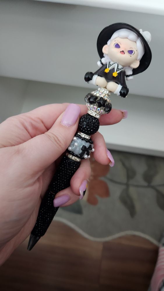 A sparkly black pen with white and black adornments at key sections in the design that all lead up to an adorable figure of a witchy little girl in a black dress, shoes, and hat who has white hair and purple eyes sitting on the top of the pen.