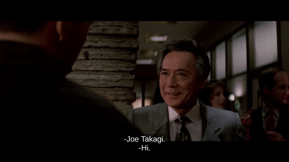 Joe Takagi (James Shigeta) introducing himself to Det. John McClane (Bruce Willis) in the classic 1988 Christmas movie Die Hard. Closed Captioning appears at the bottom of the screen reading: "Joe Takagi" followed with McClane's response of "Hi."