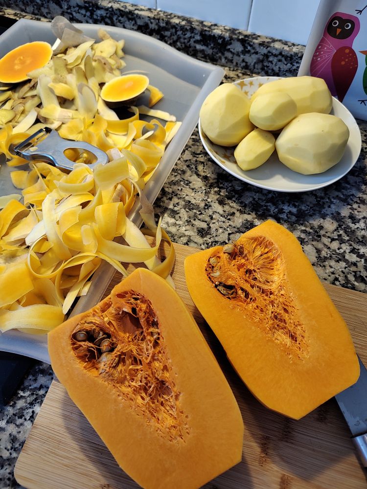 Foto de calabaza violín abierta por la mitad, la zona de las pipas llega casi hasta arriba, en un plato hay unas patatas ya peladas dispuestas a cortarse y en una bandeja tengo todas las peladuras de calabaza y patata