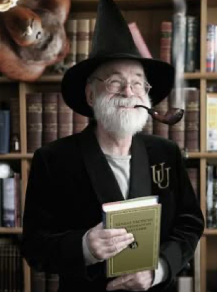 Sir Terry Pratchett, sujetando un libro mientras sonríe con una pipa en la boca y luce un elegante sombrero de bruja 