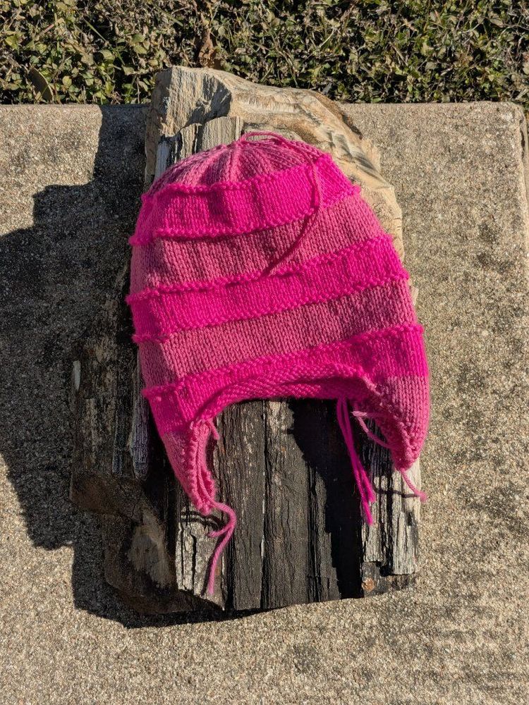 A handknitted hat is on a large piece of petrified wood.  The hat is modeled after the one worn by Edith Gru in the Despicable Me movies, with ear flaps and stripes in soft pink and VERY BRIGHT PINK.