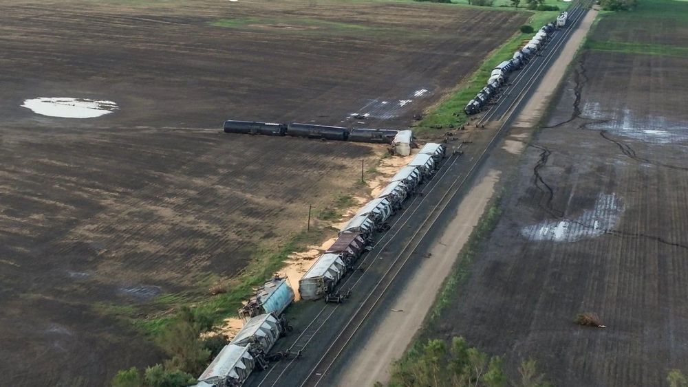 Empty fields withe a railway. A fright train composed of overturned tankers and loaded grain cars. Some of the tanker cars are hundreds of feet away from the tracks.
