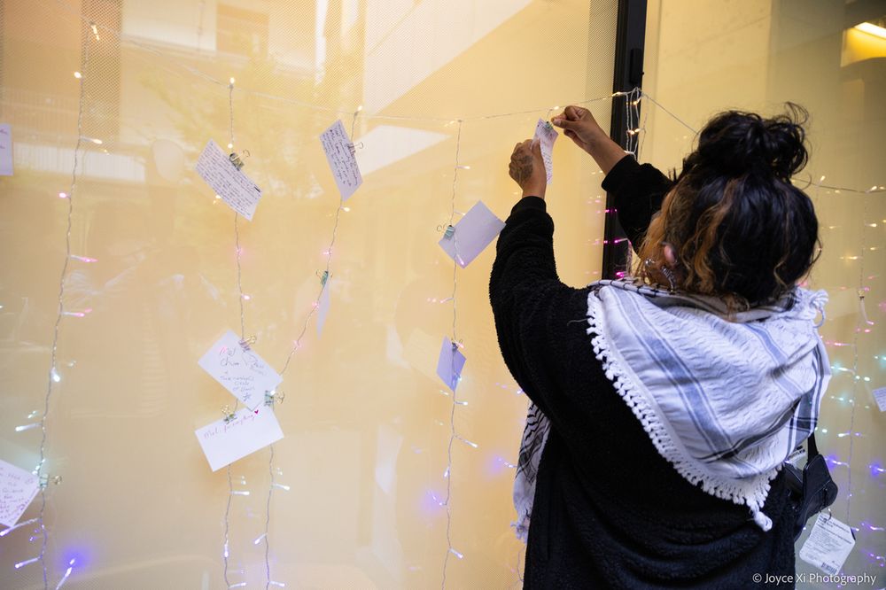A person hangs an index card on hanging fairy lights to contribute to the community art project, celebrating the people who shape disability culture. They wear a keffiyeh atop their shoulders.