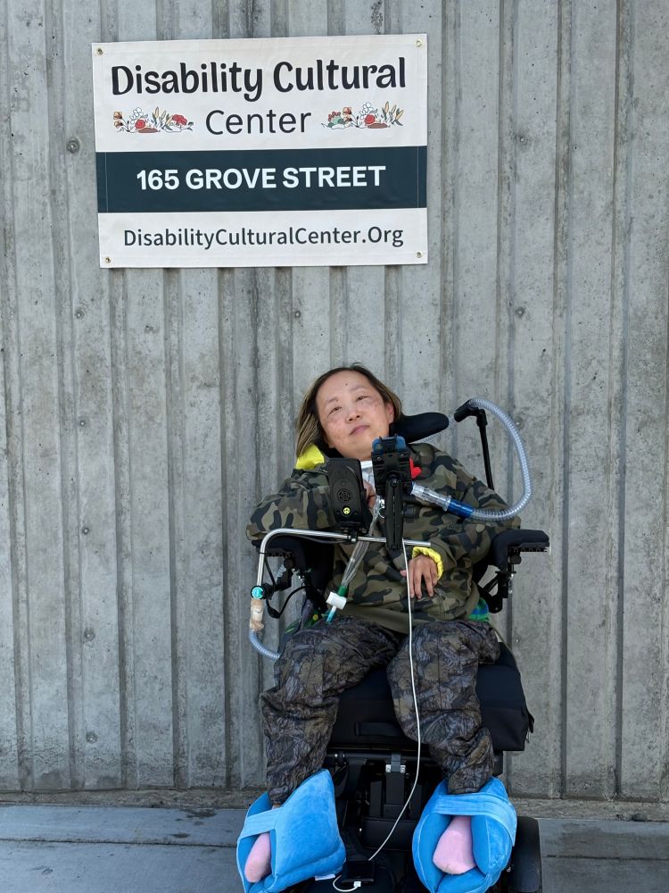 Image Description: A photo of Alice Wong, an Asian American disabled woman in a power chair, in front of the DCC sign that reads Disability Cultural Center, 165 Grove Street, www.disabilityculturalcenter.org. Alice is wearing a camouflage jacket and black pants.
