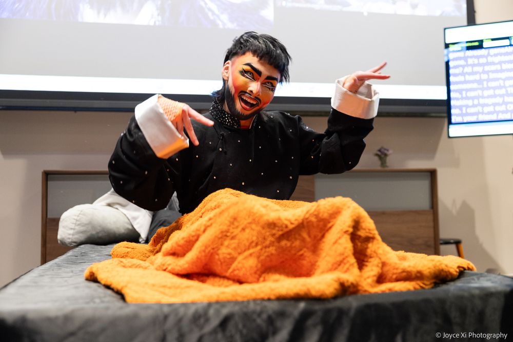 LOTUS performs from a bed with a bright orange blanket. He sits up wearing his black structured PJ top and his arms are bent and his wrists twisted to the left. He has a black mullet, a painted beard, and contacts that give his eyes a powerful look of enlarged black pupils with a neon outer ring.
